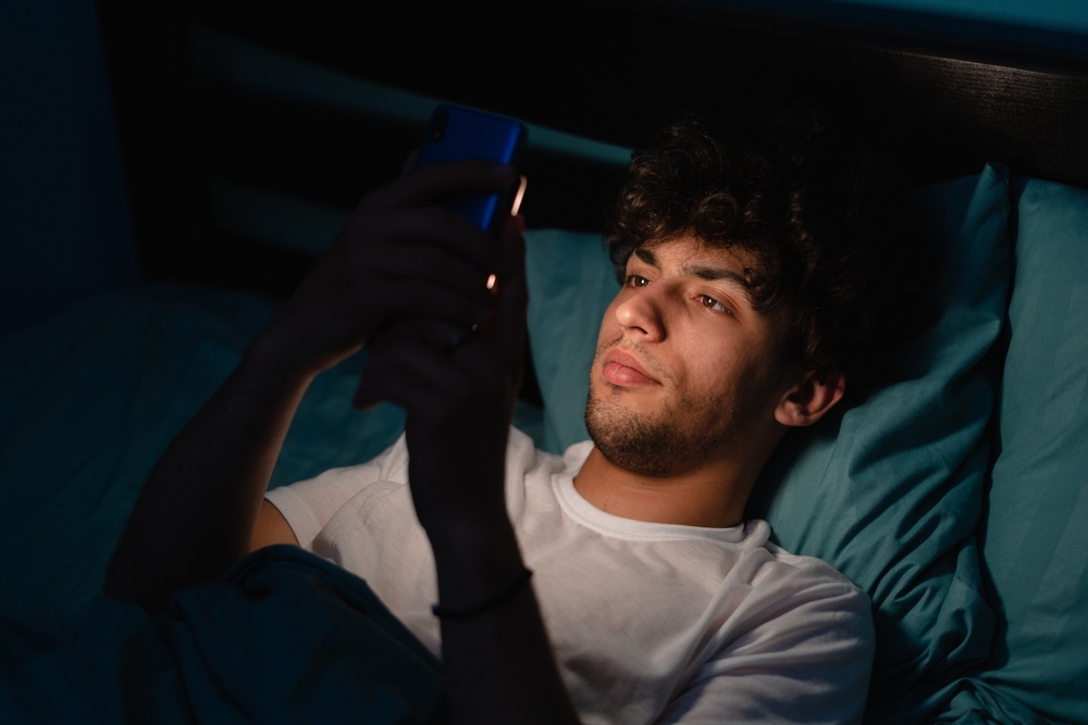 Young man lies on his bed in the dark while using his phone