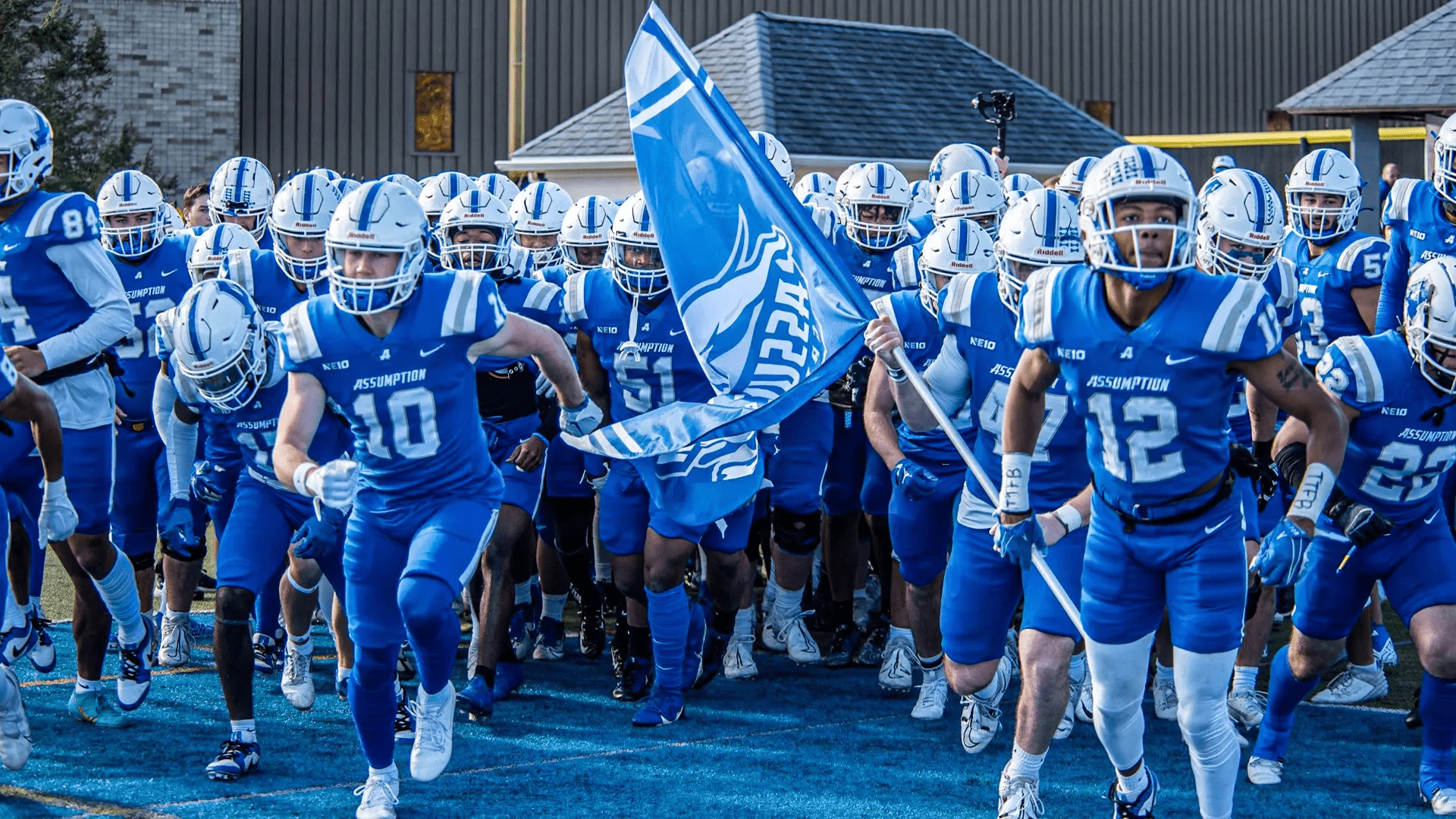 Assumption College football players running onto the football field with their school flag.